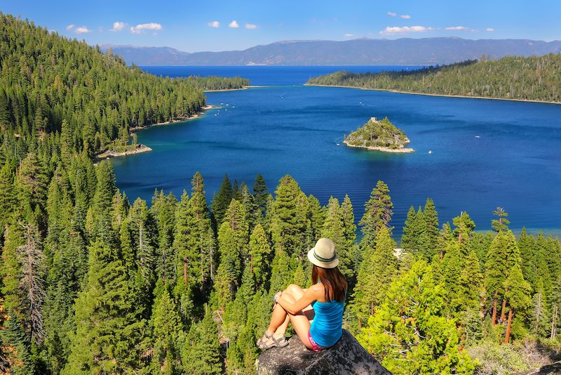 Greatest Summertime Lake Towns: Young woman enjoying the view of Emerald Bay at Lake Tahoe, California. Lake Tahoe is the largest alpine lake in North America. Photo via Shutterstock.