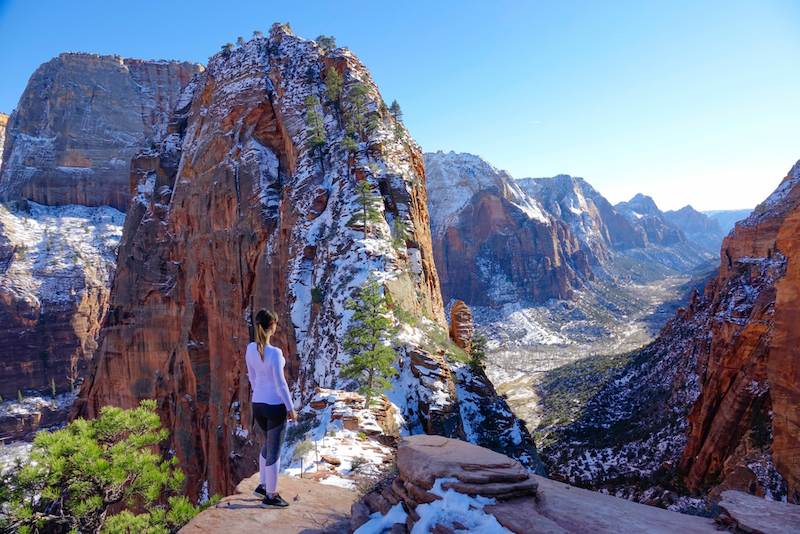 Best National Parks in Winter: Young woman observes the Angel's Landing Trail on sunny winter day under the red sandstone mountains in Zion National Park. Photo via Shutterstock.