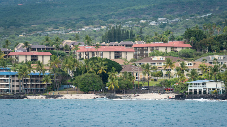 Kailua-Kona, Hawaii- JAN. 7, 2021: Oceanfront view of Honlʻs beach, next to the Kona Reef complex, known for its distinctive blue tile roof. Honlʻs is one of the few beaches in the Kona area. Photo via Shutterstock.