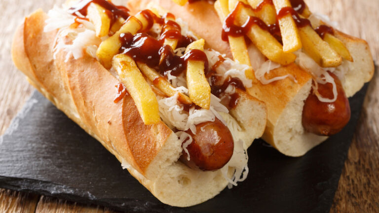 Delicious hot dog polish boy with sausage, cabbage, fries and barbecue sauce close-up. Photo via Shutterstock.